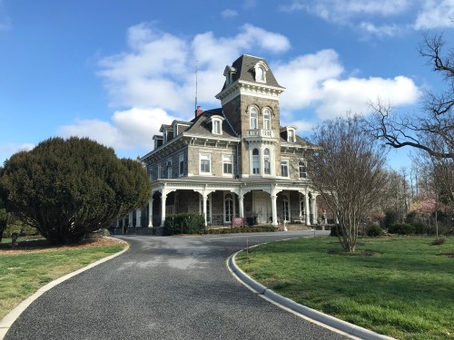 A large, historic mansion with a tower, surrounded by greenery and a gravel driveway under a blue sky.