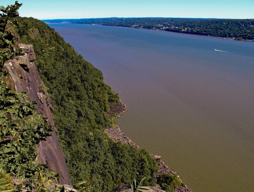 A scenic view of a river bordered by lush greenery and rocky cliffs under a clear blue sky.