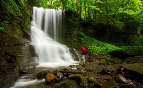 A person stands near a cascading waterfall surrounded by lush green trees and rocky terrain.