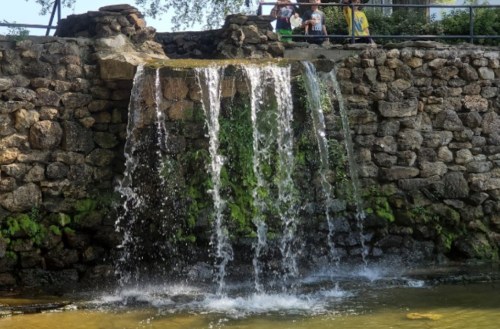 A small waterfall cascades over a stone wall, surrounded by greenery, with people visible in the background.