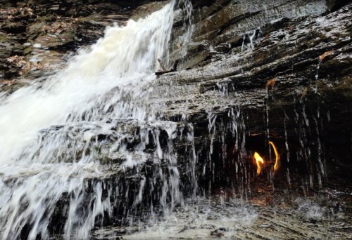 A cascading waterfall flows over rocky terrain, with water splashing and a hint of orange light visible in the background.