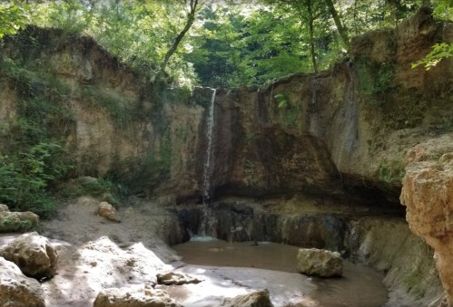 A serene waterfall cascading down rocky cliffs into a small pool, surrounded by lush green trees.