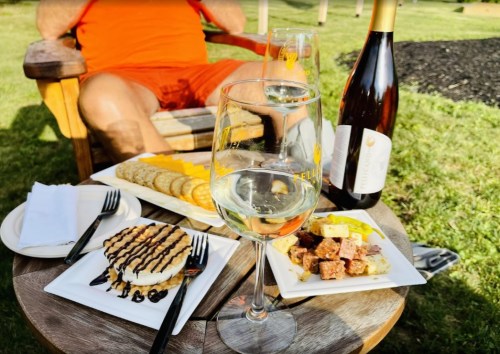 A table with a glass of white wine, cheese and crackers, dessert, and a person relaxing in the background.