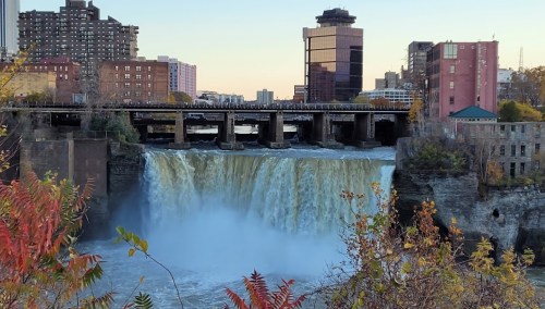 A waterfall cascades over a dam, surrounded by urban buildings and autumn foliage in a cityscape.