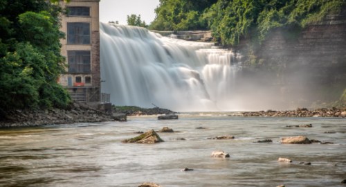 A waterfall cascades over a rocky ledge beside an old building, surrounded by lush greenery and mist.