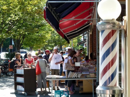 A busy street scene with people browsing books outside a shop, featuring a barber pole in the foreground.