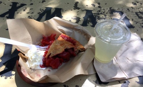 A slice of pie with strawberries and whipped cream on a paper plate, next to a clear drink in a cup with a straw.