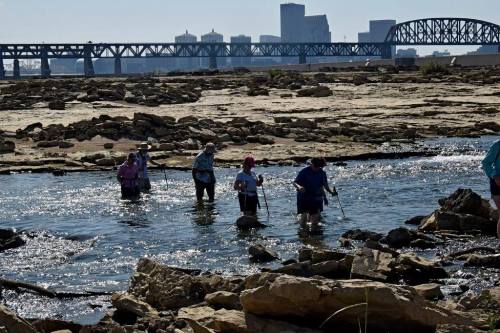 People wade through shallow water and rocks, with a city skyline and bridge visible in the background.