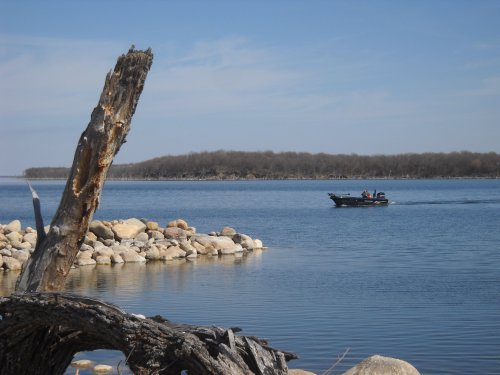 A boat glides across a calm lake, with rocky shores and a tree stump in the foreground under a clear blue sky.