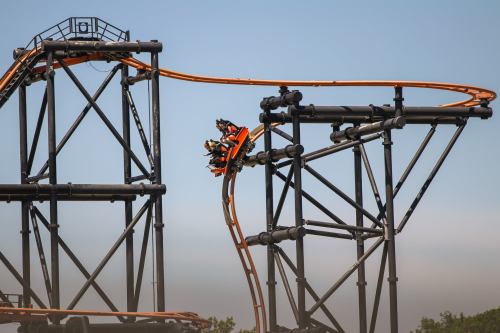 A roller coaster with an orange track twists through a black metal structure against a clear blue sky.