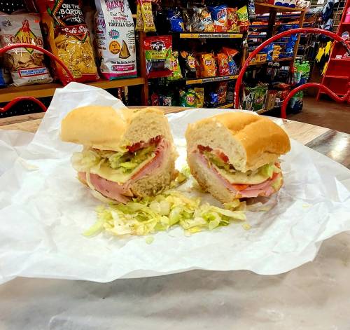 A sandwich with layers of ham, lettuce, and tomato, cut in half, on a white paper in a colorful store setting.