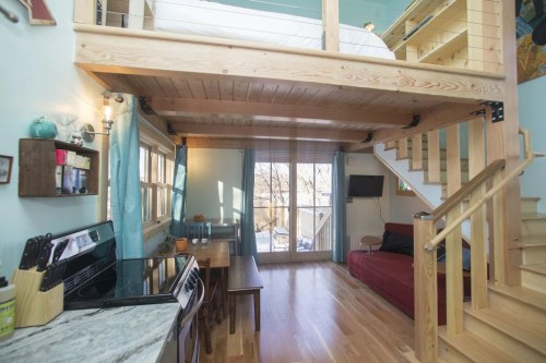 Cozy interior of a small home featuring a lofted bed, kitchen, dining area, and large windows with natural light.