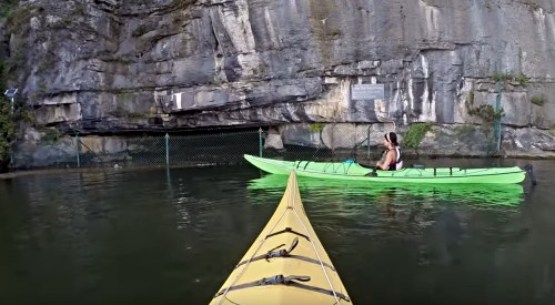 A yellow kayak in the foreground with a green kayak nearby, surrounded by rocky cliffs and calm water.