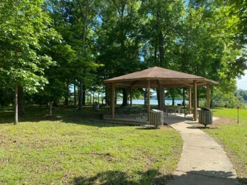 A shaded gazebo surrounded by trees, with a path leading to a lake in the background on a sunny day.