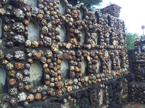 A textured wall made of colorful stones and shells, featuring arched niches and a natural, rustic appearance.