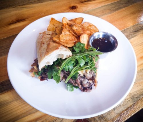 A plate featuring a sandwich with beef, greens, and cheese, accompanied by crispy potato chips and a dipping sauce.