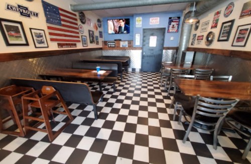 A diner interior with checkered floor, wooden tables, and vintage car memorabilia on the walls. A TV is visible.