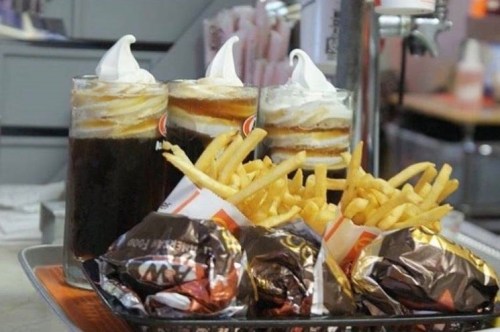 Three glasses of root beer floats with whipped cream, alongside a tray of crispy French fries and wrapped burgers.