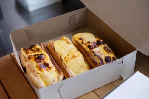 Three toasted hot dog buns filled with melted cheese, placed in a brown takeout box.