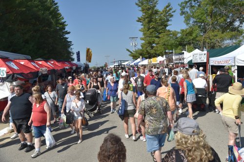 A busy outdoor market with crowds of people walking between colorful vendor tents under a clear blue sky.