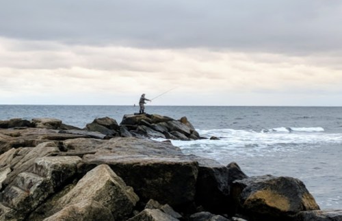 A lone fisherman stands on rocky shore, casting a line into the calm ocean under a cloudy sky.