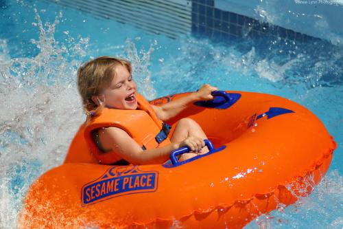 A joyful child in an orange inflatable tube splashes through water at a water park, wearing a life jacket.