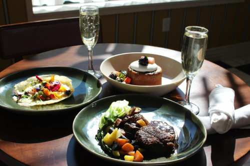 A table set with three gourmet dishes, sparkling drinks, and a napkin, illuminated by natural light.
