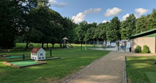 A grassy park with a mini-golf area, playground, and basketball court under a blue sky with scattered clouds.