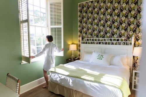 A person in a white robe stands by a window in a stylish bedroom with green walls and floral wallpaper.