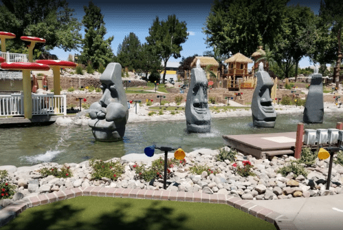 A mini-golf course featuring large stone sculptures, water features, and lush greenery in a sunny outdoor setting.