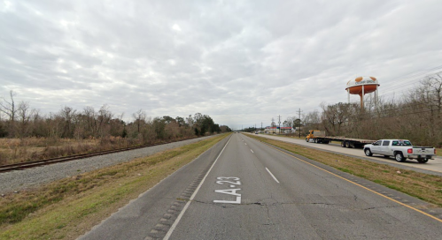 A wide road with a highway sign for LA-23, alongside a railway and a water tower in a rural area under a cloudy sky.