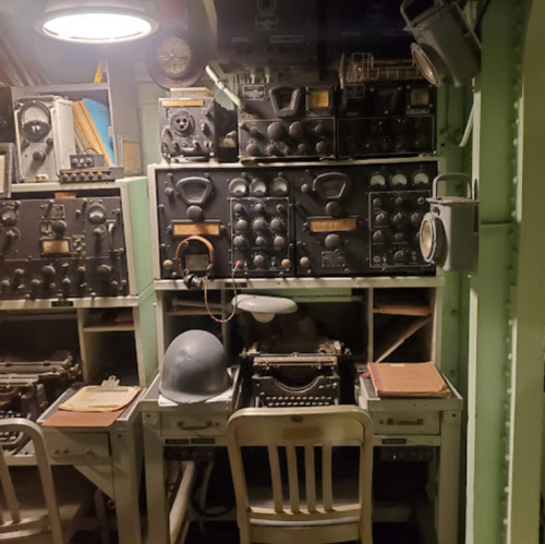 A vintage communication room with old radios, a typewriter, and a helmet on a desk, surrounded by green walls.