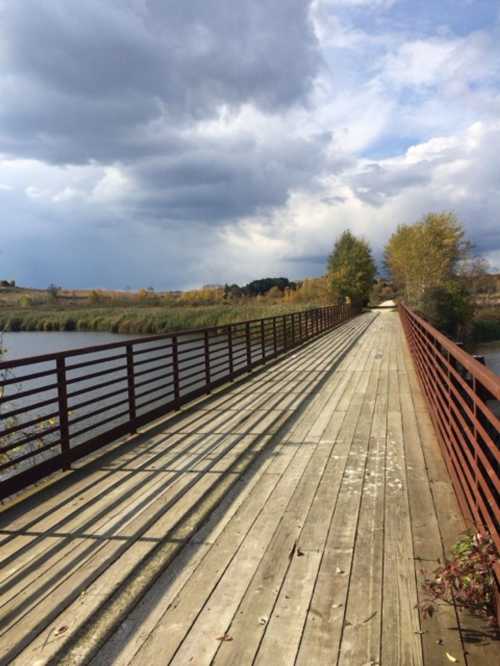 A wooden bridge stretches over water, lined with trees, under a cloudy sky.