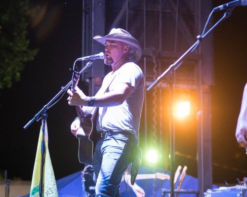 A musician in a cowboy hat performs on stage with an acoustic guitar, illuminated by stage lights at night.