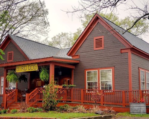 A charming gray house with red trim and a sign reading "Bird Dog," surrounded by greenery and a welcoming porch.