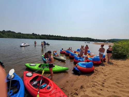 A sunny beach scene with people kayaking and relaxing by the water, surrounded by colorful kayaks and greenery.