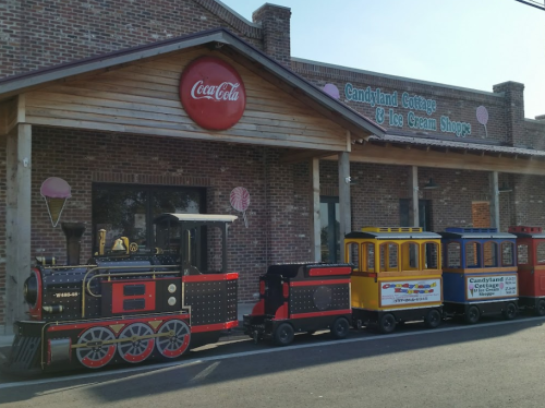 A colorful miniature train parked outside Candyland Cottage & Ice Cream Shoppe, featuring a Coca-Cola sign.