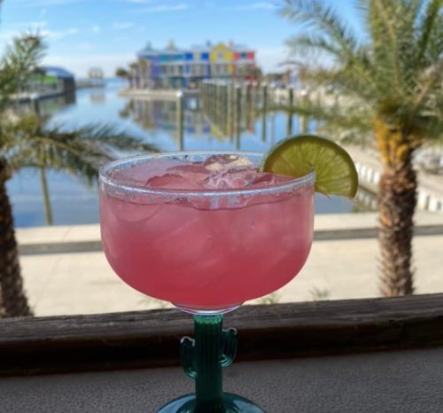 A vibrant pink cocktail with a lime slice, set against a scenic waterfront view with colorful buildings and palm trees.