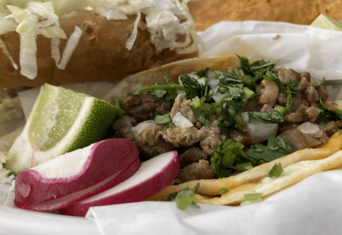 A close-up of a taco filled with meat, topped with onions and cilantro, served with lime and radish slices.