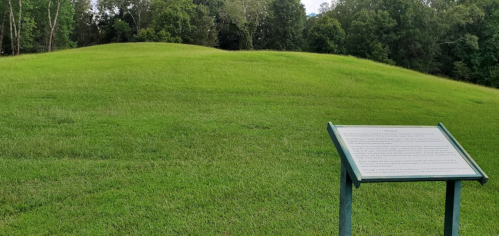 A grassy hill with a historical information sign in the foreground, surrounded by trees.