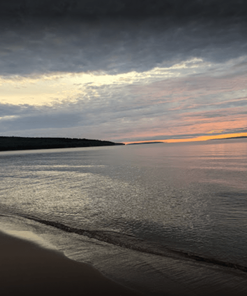 A serene beach scene at sunset, with calm waters reflecting colorful clouds and a distant shoreline.