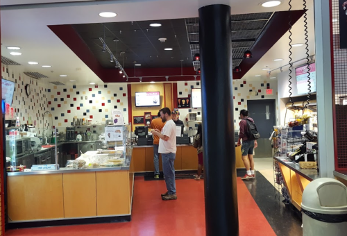 A busy café interior with customers ordering at the counter and a variety of food displays. Red and black decor.