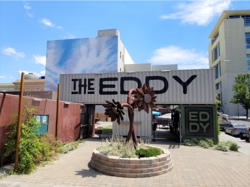 Entrance to The Eddy, featuring a large sign and a flower sculpture, with a blue sky and buildings in the background.