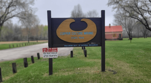Sign for Copper Culture Mound State Park, with a red building in the background and a pet policy notice nearby.