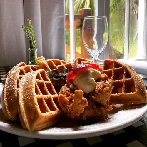 A plate of waffles with fried chicken, topped with a strawberry and a dollop of cream, beside a glass and a small vase.