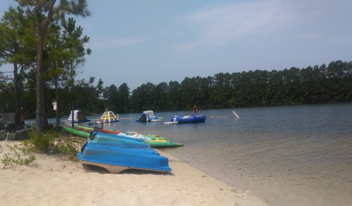 A sandy beach with colorful kayaks, inflatable water toys, and a calm lake surrounded by trees under a clear sky.