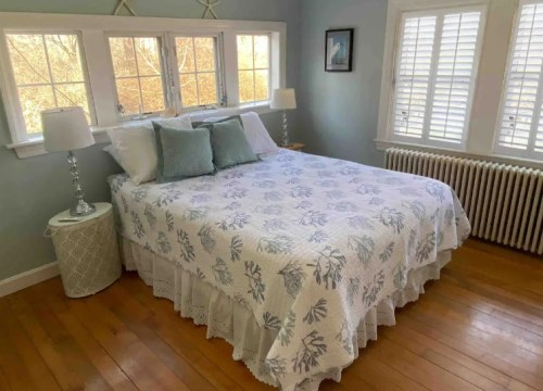 Cozy bedroom with a double bed, light blue walls, and large windows, featuring a floral quilt and wooden floors.