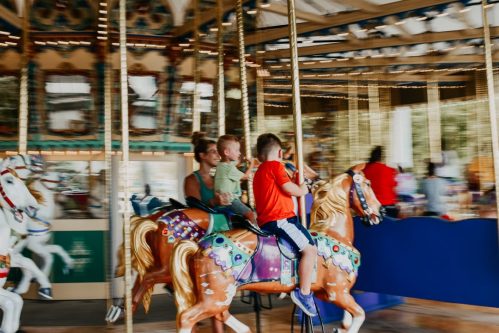 A child rides a colorful carousel horse with a woman beside him, both smiling in a lively amusement park setting.