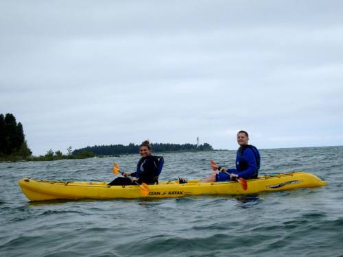 Two people in blue wetsuits sit in a yellow kayak on a calm body of water, with a cloudy sky in the background.