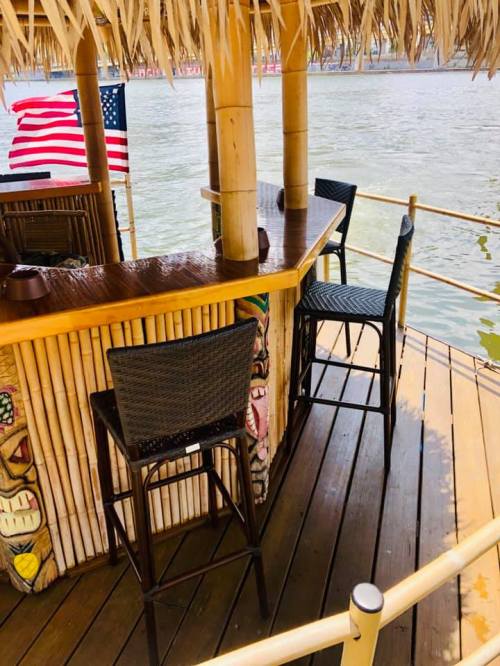 A bamboo bar with high chairs by the water, featuring a thatched roof and an American flag in the background.